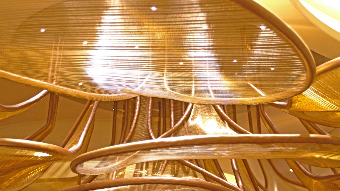Chandelier monumental en maille métallique – Celebrity Xcel - <p>Chandelier monumental en maille métallique dorée installé dans le restaurant Le Voyage à bord du Celebrity Xcel. Cette réalisation met en valeur la transparence et les reflets de la maille métallique dans un projet d’architecture intérieure haut de gamme.</p>